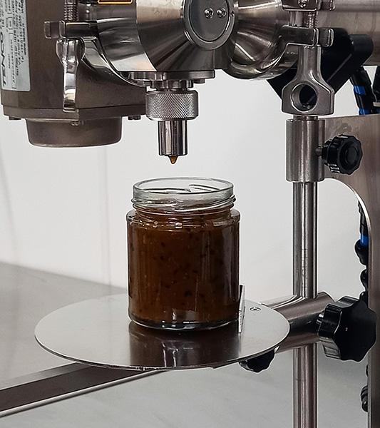 A close up of a filling machine filling a glass jar with mango chutney.