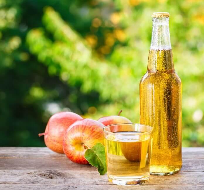 A glass cider bottle, glass and apples on a table, with a green background.
