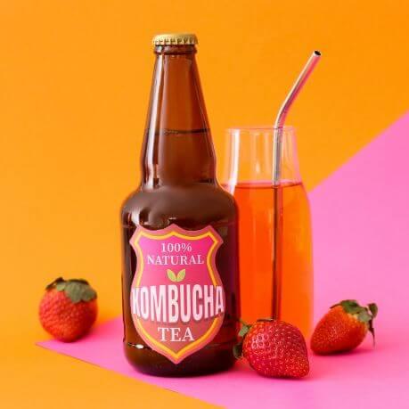 A glass kombucha bottle, with a glass and straw and two strawberries next to it, on a pink and orange background.